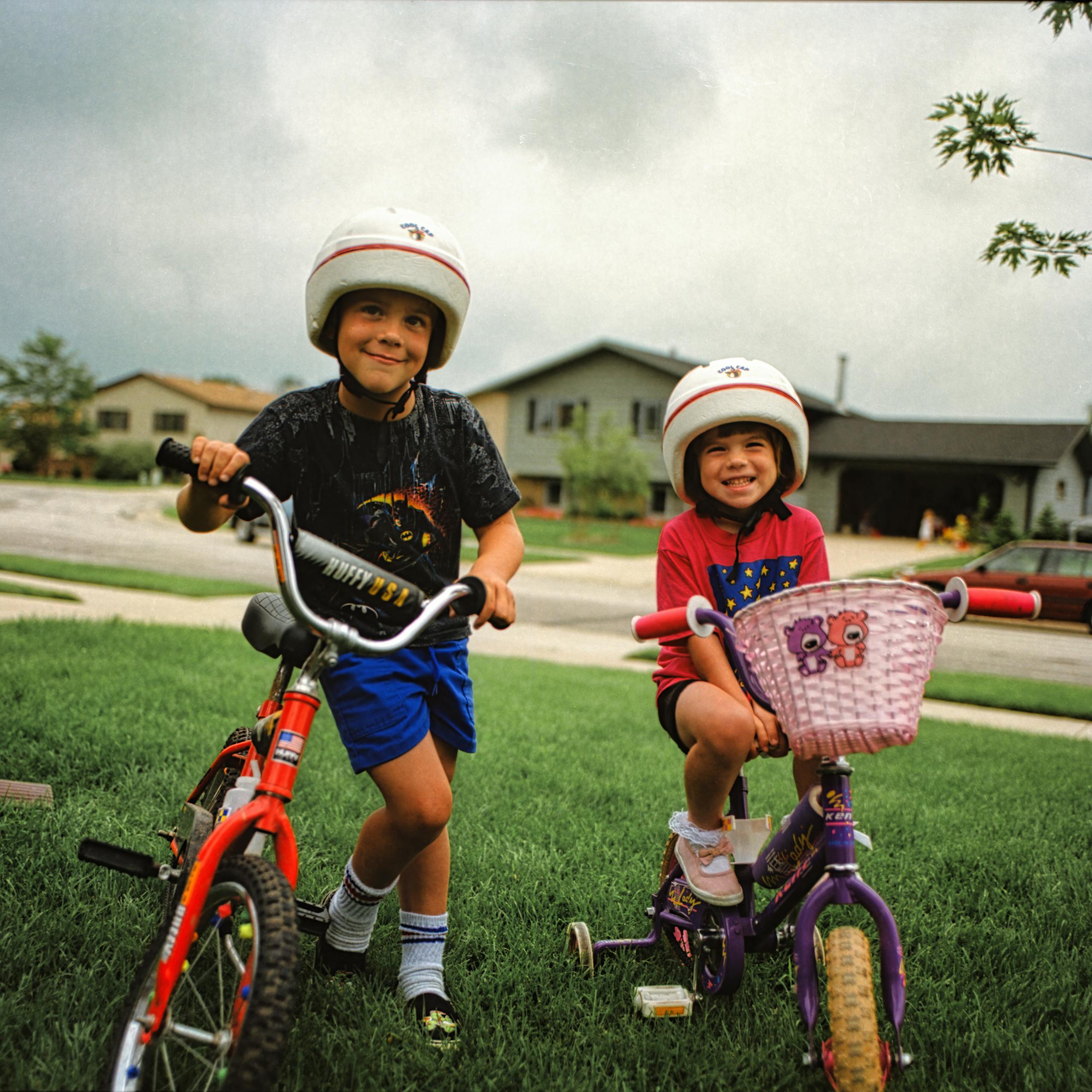 Smiling kids riding bikes with helmets in a suburban neighborhood on a cloudy day.